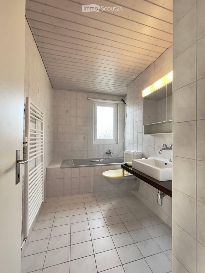 Bathroom with bathtub, toilet, sink, and mirror, white tiles, wooden ceiling, and a window.