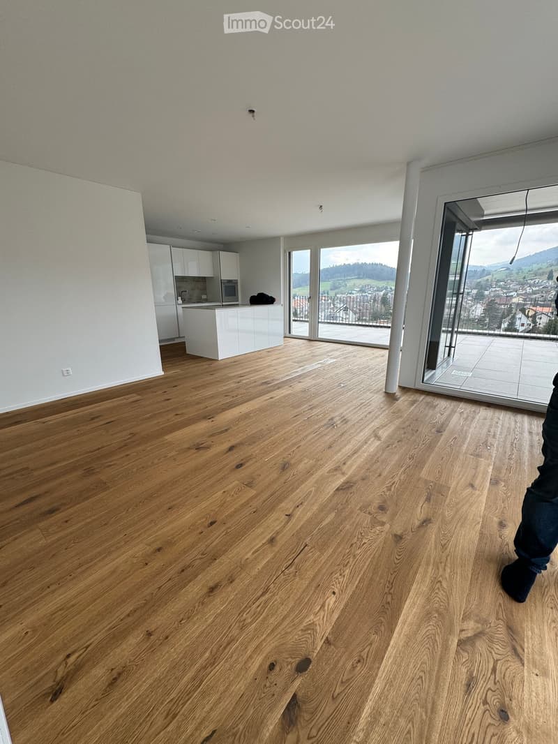 Open-plan living and kitchen area with light wooden flooring, white kitchen cabinets, and a scenic view through large windows and sliding doors.
