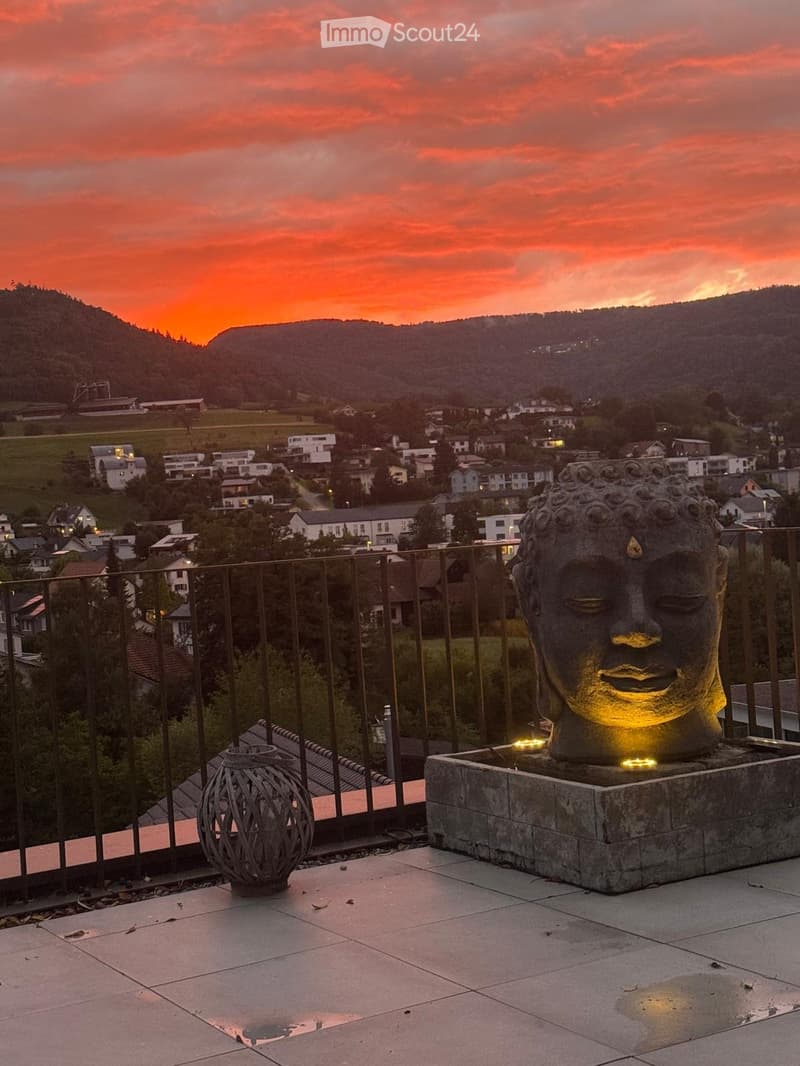 Outdoor area with tiled floor, metal railing, Buddha statue, small lantern, sunset backdrop with mountains and village.