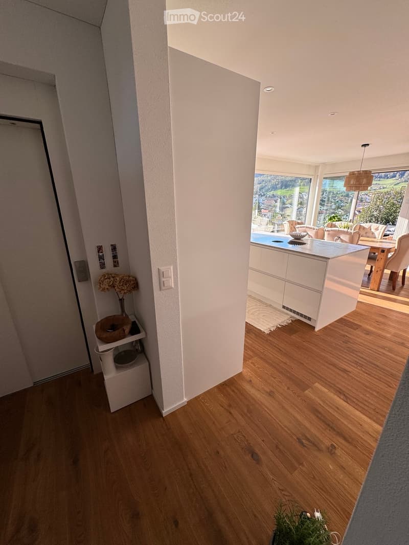 Open-plan kitchen and living area with wooden flooring, white cabinetry, and large windows overlooking a scenic view.