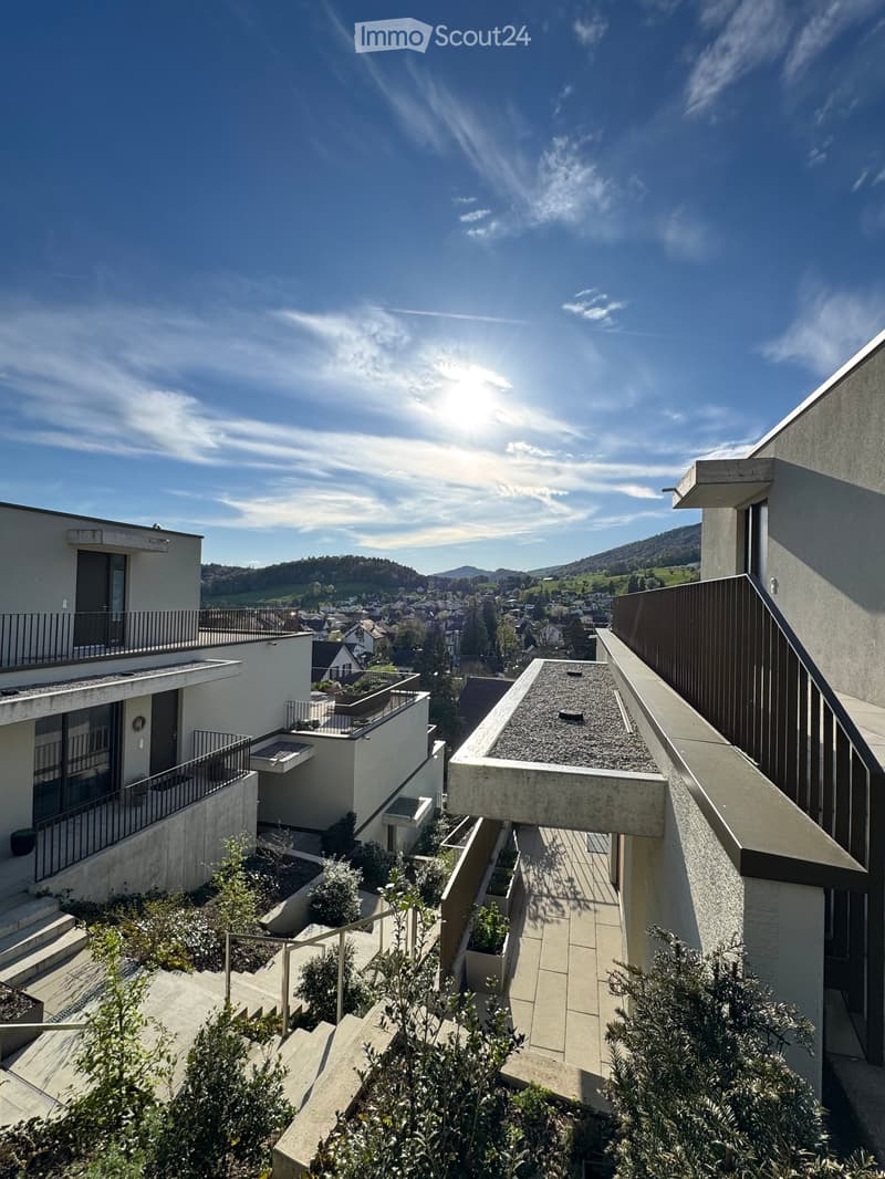 Modern building with multiple balconies, concrete and glass design, surrounded by greenery, elevated view of the town and mountains