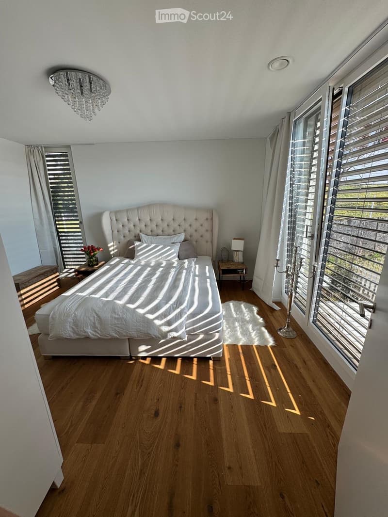 Bedroom with wooden floor, white walls, large bed, chandelier, nightstands, and large windows with curtains