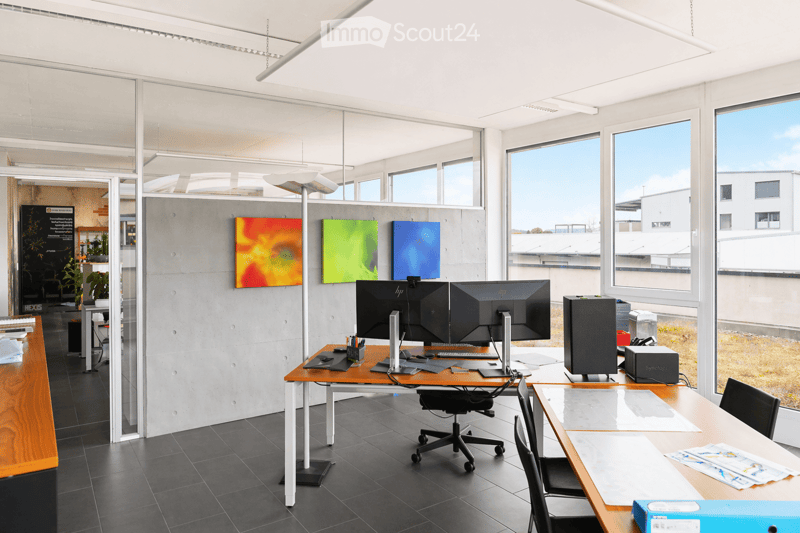 Modern office space with large windows, desks, chairs, computer monitors, keyboards, mice, speakers, and artworks on the walls.