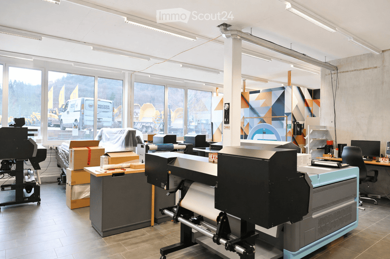 Modern office space with large windows, industrial-style decor, and various equipment including printers, tables, and chairs.