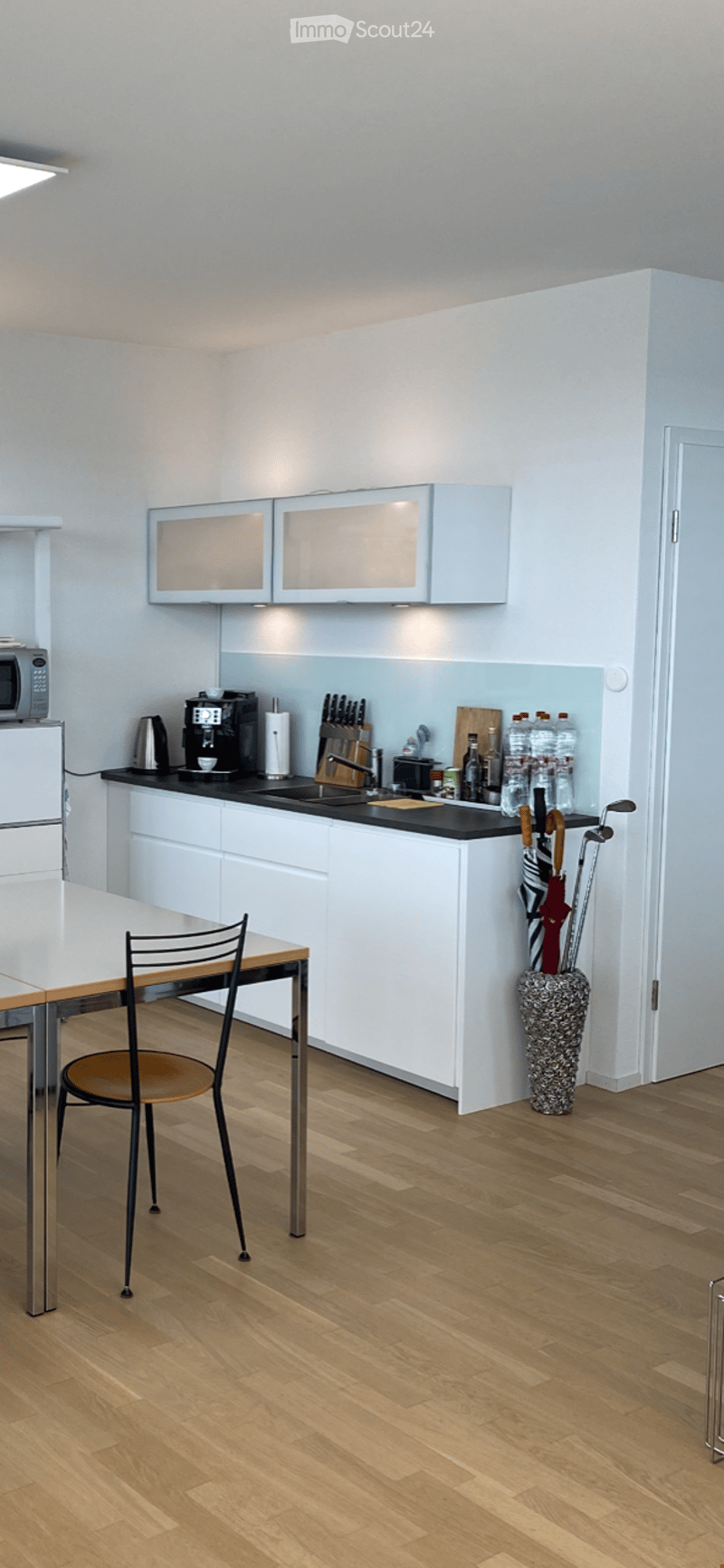 Modern kitchen with white cabinets, black countertops, stainless steel appliances, and a small dining area with a table and chair. Wooden flooring and a decorative umbrella stand are also visible.