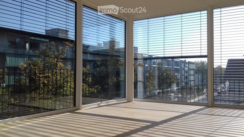 Empty room with large windows and blinds, overlooking a cityscape with buildings and trees. The floor is light-colored, and the space is well-lit with natural light.