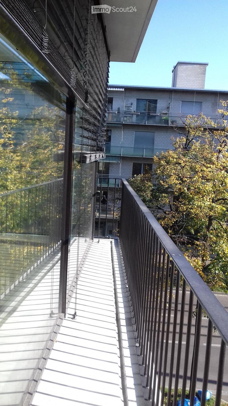 Modern balcony with glass railing, metal balustrade, and steps leading to a rooftop area. The building in the background has a contemporary design with large windows and a balcony.