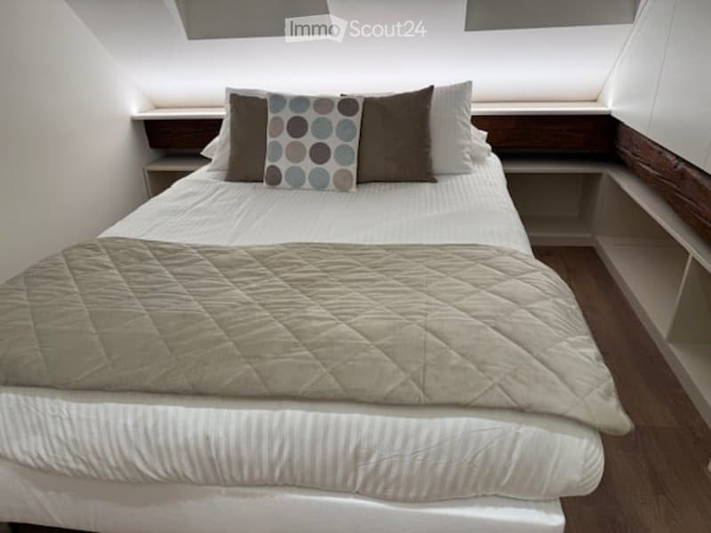 A cozy bedroom with a neatly made bed, featuring a quilted beige duvet, white sheets, and decorative pillows, including a patterned pillow and two brown pillows. The room has built-in wooden shelves on either side of the bed and a slanted ceiling with rec