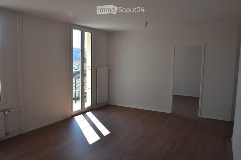 Empty room with wooden flooring, large window with view, radiator, and a doorway leading to another space.