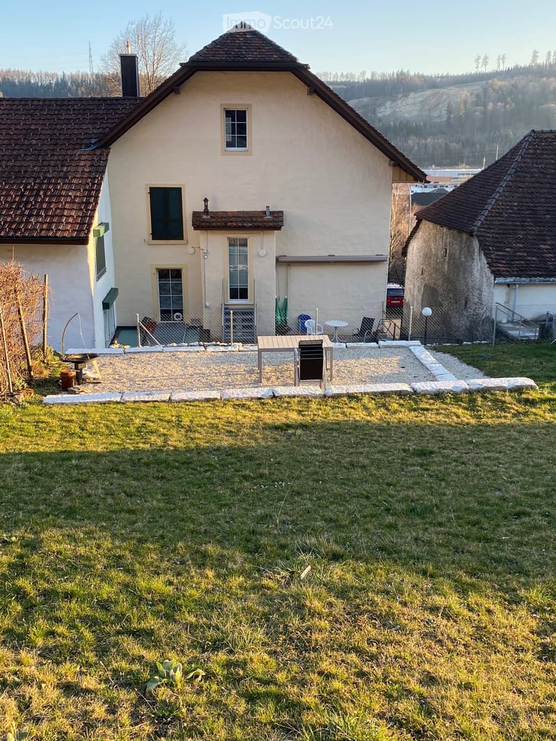 A two-story house with a tiled roof, surrounded by a grassy lawn and a small patio area with outdoor furniture.