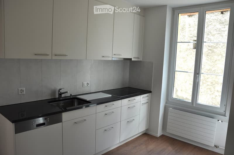 Modern kitchen with white cabinets, black countertop, stainless steel sink, and dishwasher. Features a window with a view of a stone wall outside.