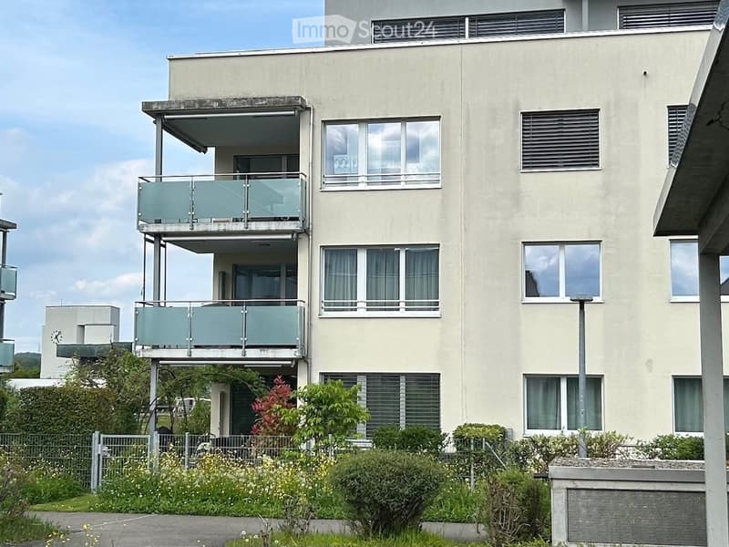 Modern multi-story building with balconies, large windows, and surrounding greenery.