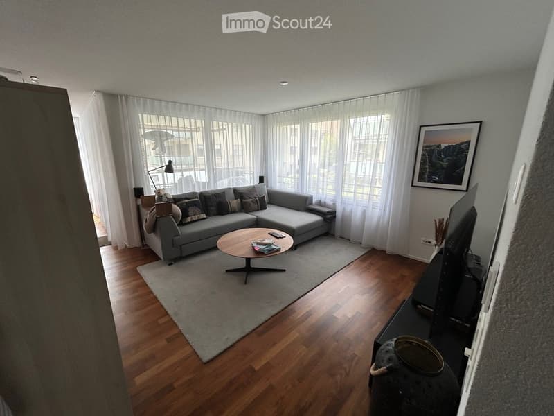 Modern living room with a grey sectional sofa, wooden coffee table, floor lamp, and large windows with white curtains.
