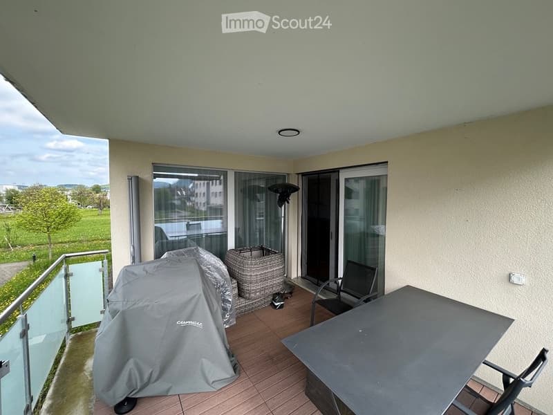 Balcony with wooden decking, glass sliding doors, covered grill, wicker basket, table, and chairs.