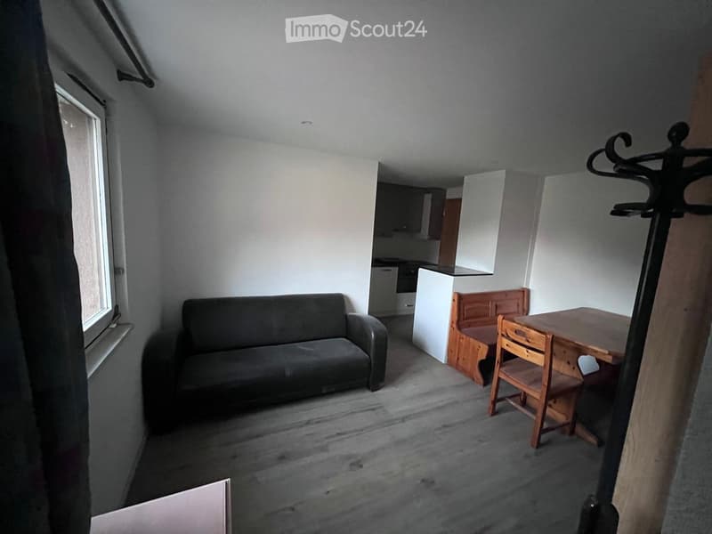 an empty room with a sofa, a desk with two chairs, a window, and a kitchen in the background