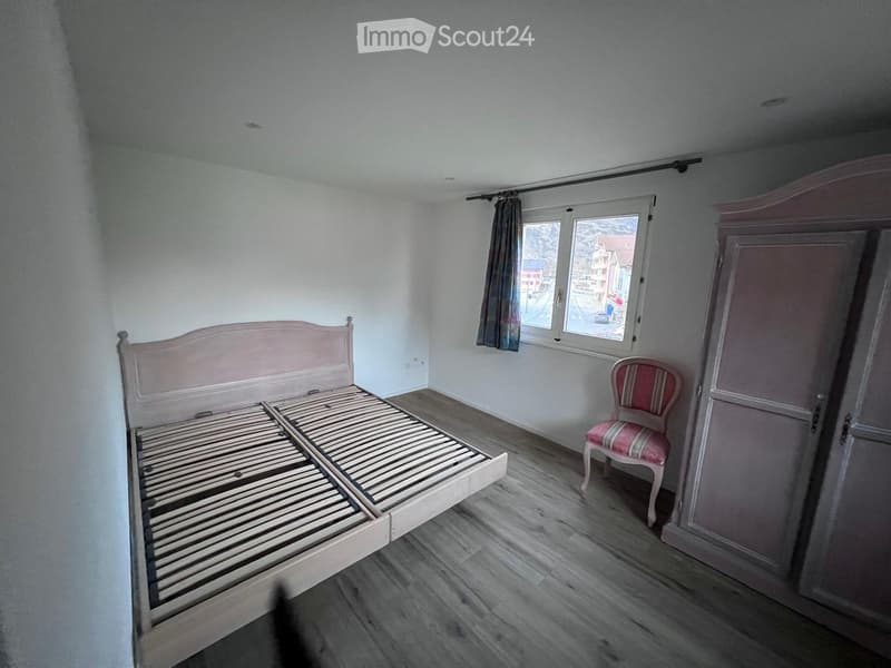 room with wooden bed, wooden floor, white walls, window with view of town, pink chair, and wardrobe