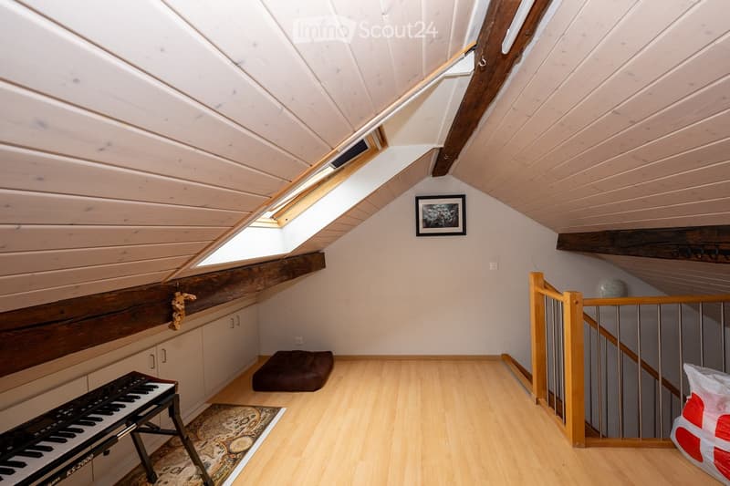 Bright attic room with wooden ceiling, skylight windows, and a staircase leading upwards. Contains a small keyboard on a stand, a brown stool, a decorative rug, and a picture frame on the wall.
