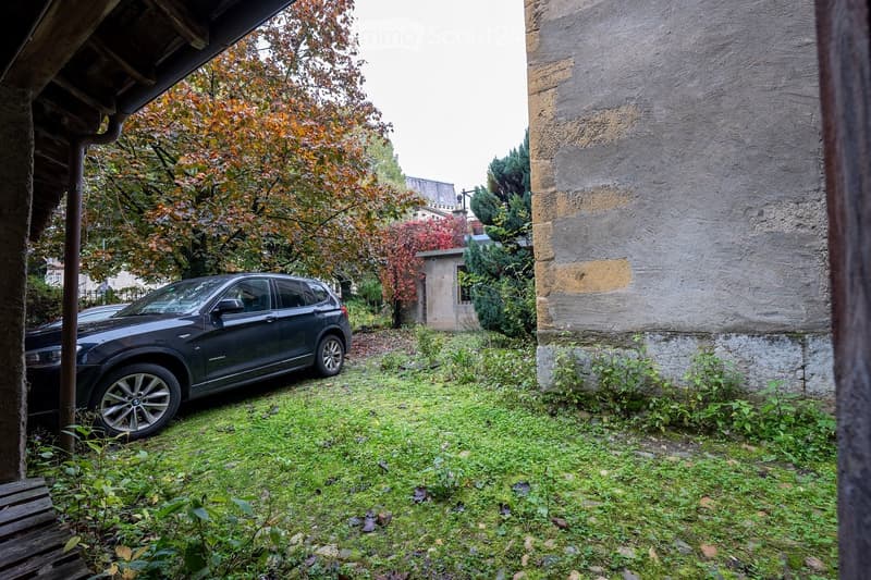 A grassy outdoor area with a parked SUV, surrounded by trees and a stone wall. The area is partially covered by a wooden structure on the left side.