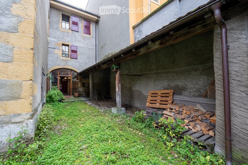 Courtyard with stone walls, wooden bench, stack of firewood, small garden area with grass and plants, and an arched entrance to a building.