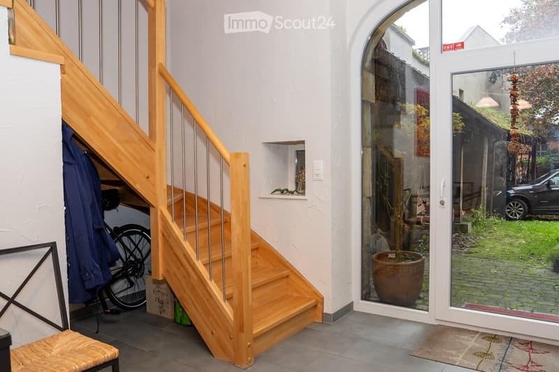 Modern hallway with wooden staircase, glass door leading to outdoor area, bicycle, coats, and potted plant visible.