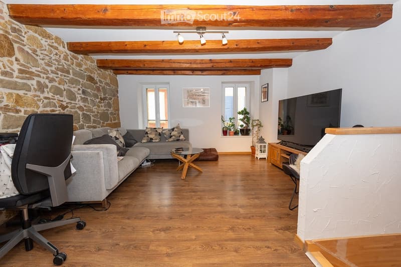 Living room with exposed wooden beams, stone wall, grey sofa, coffee table, TV, and potted plants on windowsill.