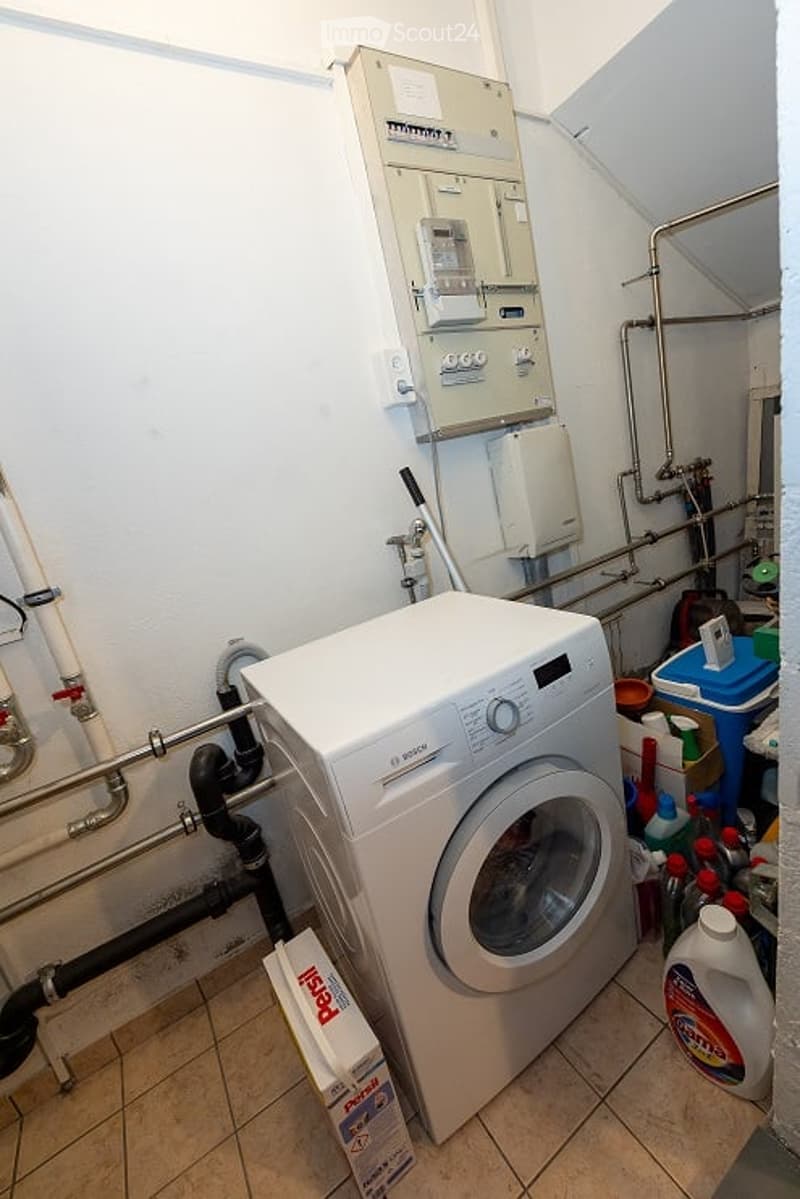 Utility room with a white washing machine, cleaning supplies, and exposed pipes.
