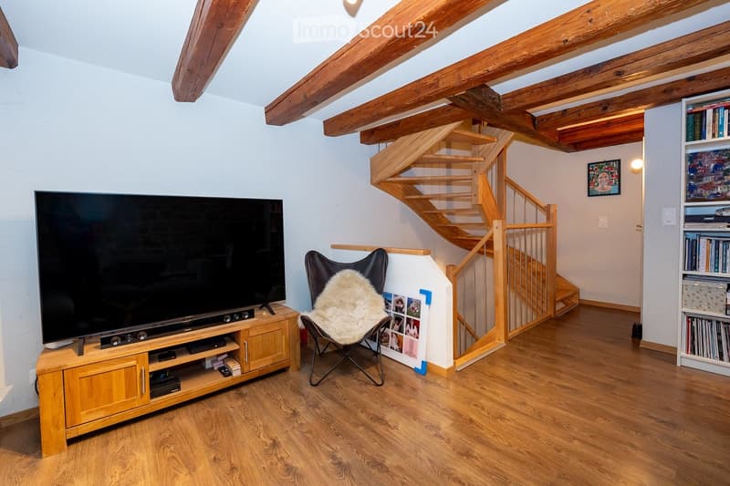Living room with wooden floor, exposed wooden beams, television on wooden cabinet, a chair with a fur throw, and a wooden staircase leading upwards.