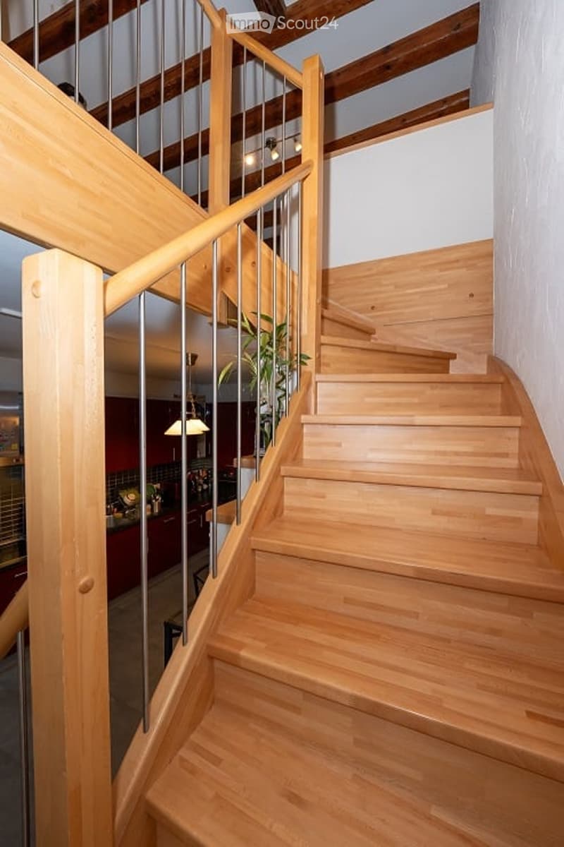 Spacious wooden staircase with a modern metal railing, leading to an upper level. The staircase features light-colored wooden steps and a polished finish, with exposed wooden beams on the ceiling. A small potted plant is placed near the base of the stairs