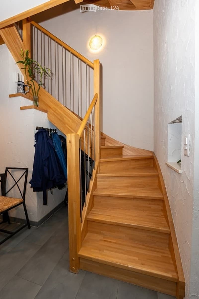 Wooden staircase with metal railing, coat rack with jackets, small plant on shelf, and a chair near the base.