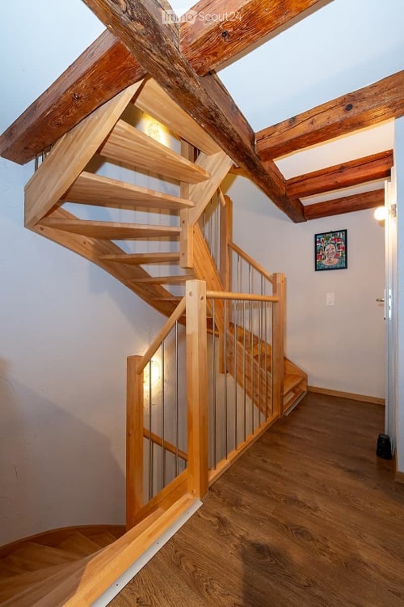 Spacious hallway with wooden flooring and exposed wooden beams on the ceiling. A modern wooden staircase with a metal railing ascends to an upper level. A small framed artwork hangs on the wall.