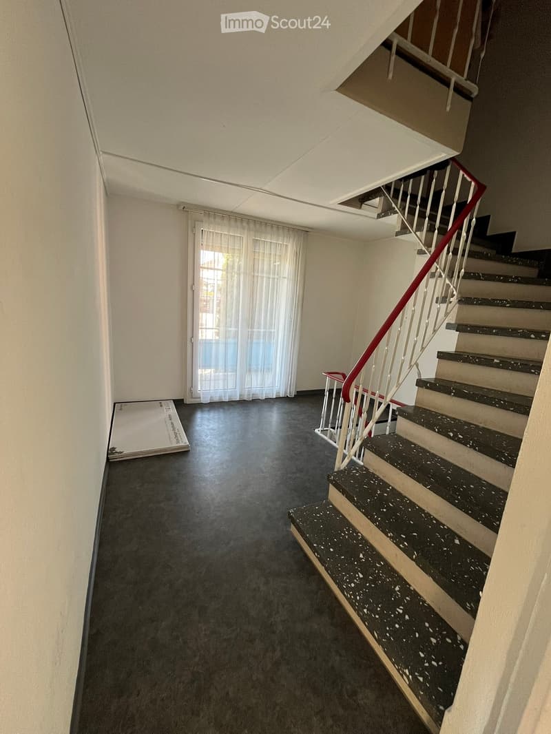 Empty hallway with a staircase leading upwards, featuring a dark tiled floor and white walls. A glass door with white curtains is visible on the left side.