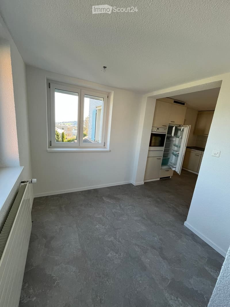 Empty room with grey carpeted floor, white walls, a window with white frame, radiator, and an open doorway leading to a kitchen area with cabinets and appliances.