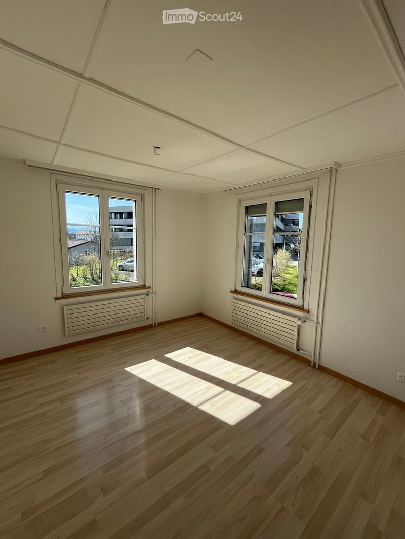 Empty room with wooden flooring, white walls, and large windows with radiators beneath them. Sunlight streams in, casting shadows on the floor.