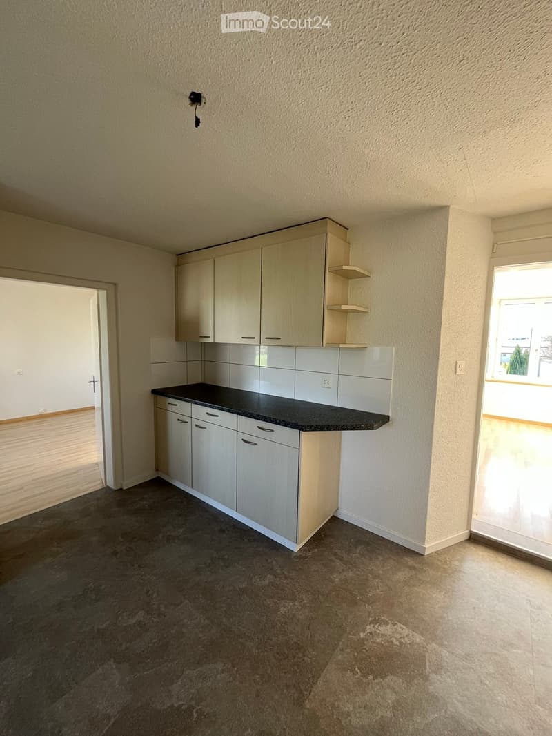 Empty kitchen with light-colored cabinets, black countertop, and tiled backsplash. The space includes a doorway and a window letting in natural light.