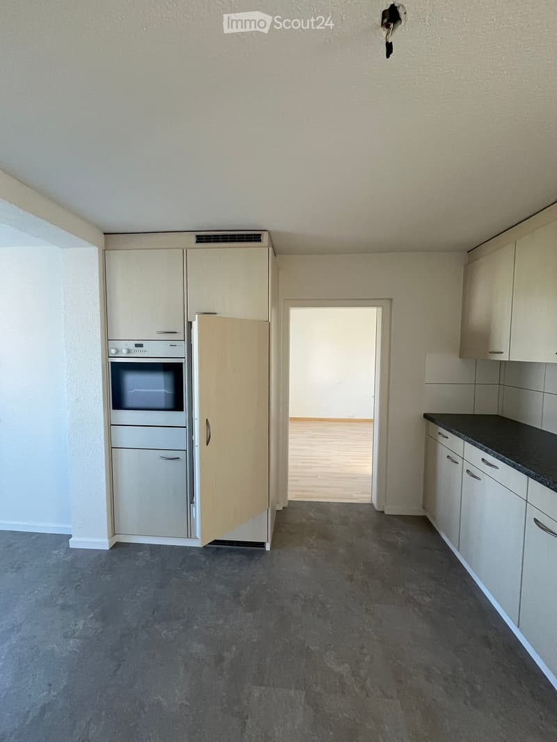 Empty kitchen with light-colored cabinets, a built-in oven, and a dark countertop. The floor is gray concrete, and the walls are light beige.