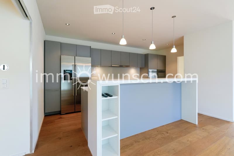 Modern kitchen with grey cabinets, stainless steel refrigerator, white counter, and wooden flooring.