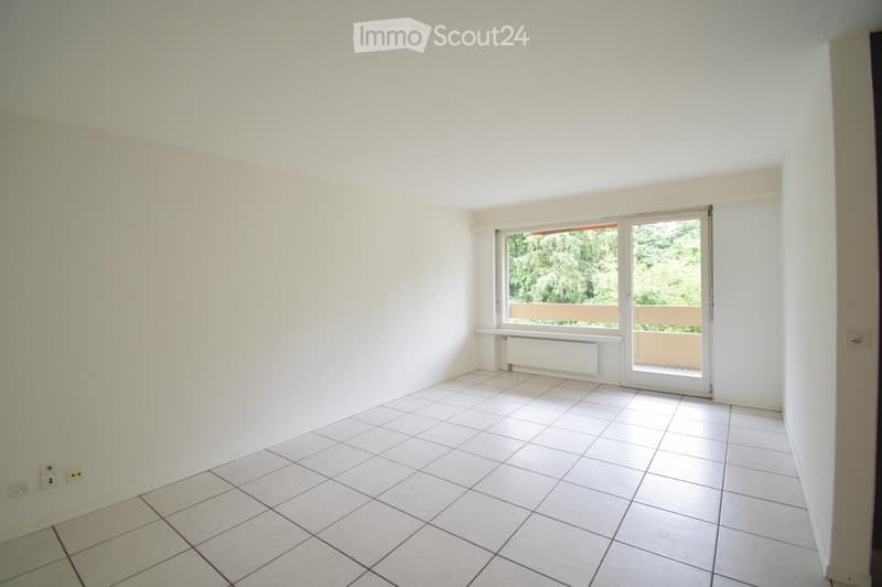 Empty room with white tiled flooring, white walls, and a large window with a sliding door leading to a balcony. There is a radiator below the window and electrical outlets on the walls.