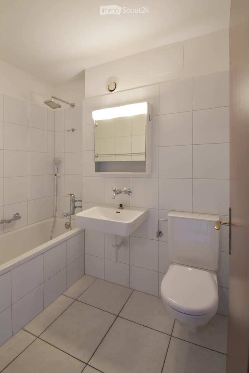 Modern bathroom with white tiles, bathtub, sink, toilet, and illuminated mirror.