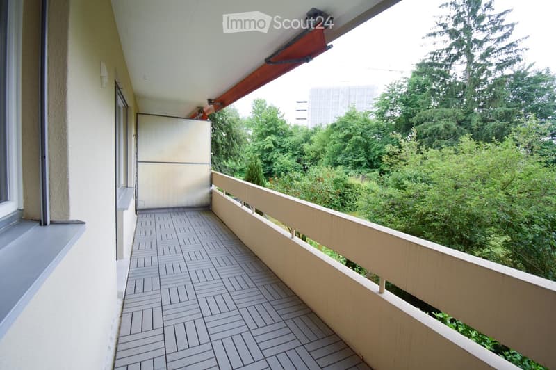 Balcony with tiled floor, wooden railing, and a view of greenery and trees.