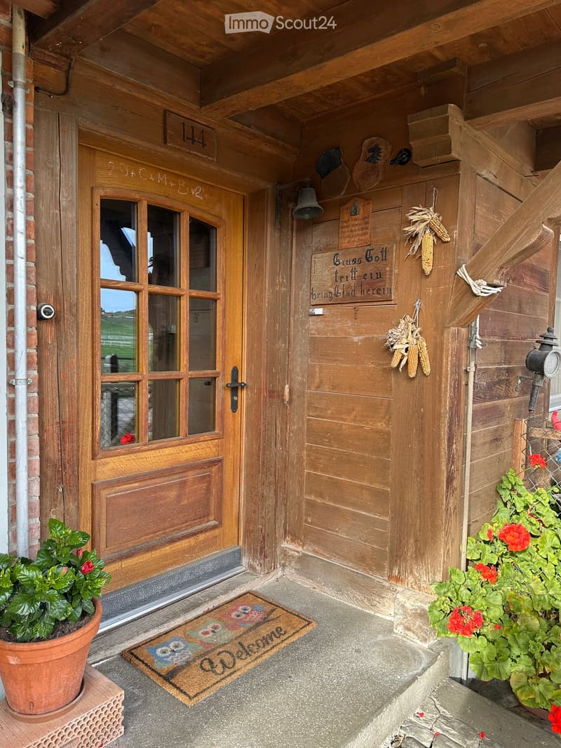 Wooden entrance with welcome mat, two potted plants, door with glass panels, sign 144, metal bell