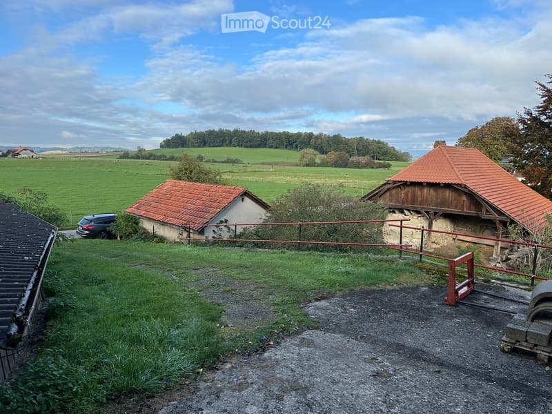two houses, one with red roof, one with white roof, fenced off, car parked outside, fields in the background