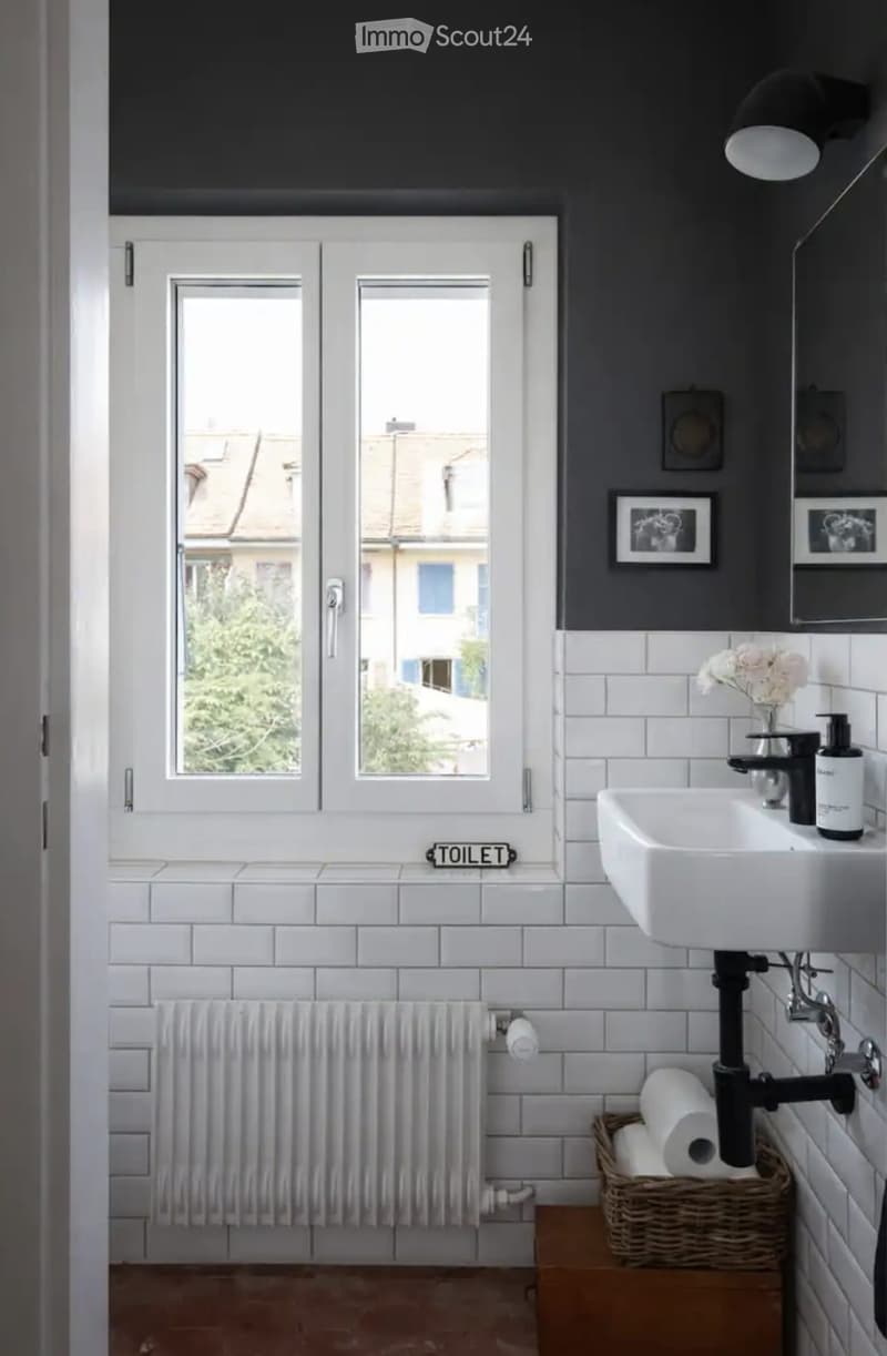 Modern bathroom with white tiles, dark grey upper walls, a white sink with a black faucet, a radiator, a toilet paper holder, and a wicker basket with toilet paper rolls.