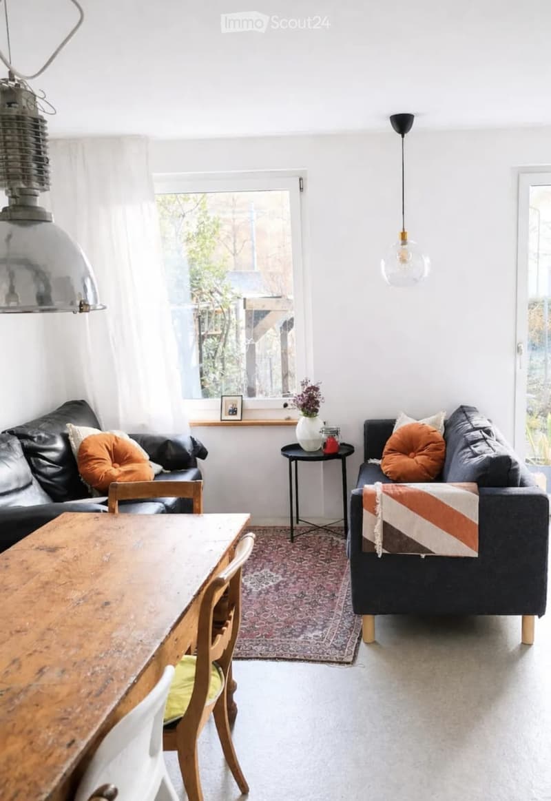 Bright living room with two sofas, a wooden dining table, chairs, a small side table, and a decorative vase on the windowsill.