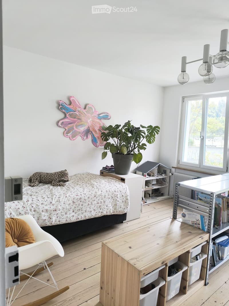 Bright bedroom with wooden flooring, a bed with a patterned duvet, a white rocking chair, a wooden table with storage bins, a shelf with books and items, a large potted plant, and a colorful wall decoration.