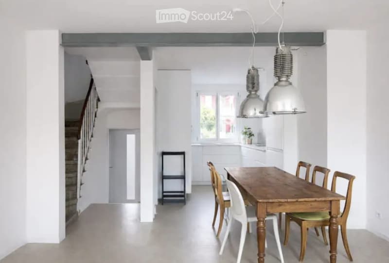 Bright, open-plan dining area with a wooden dining table, white and wooden chairs, pendant lights, and a small stool near the kitchen area.