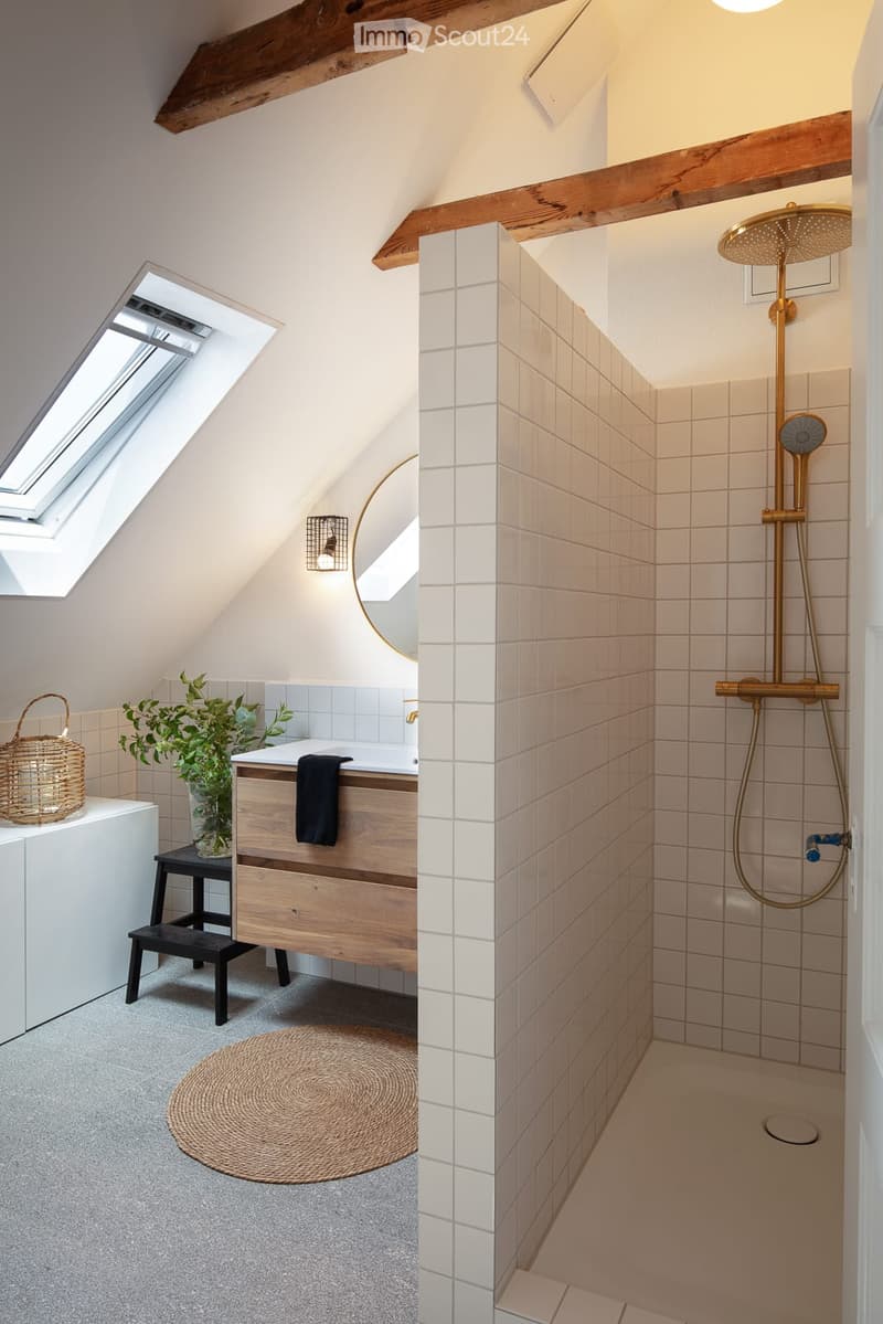Modern bathroom with a wooden vanity, white tiles, a large shower with gold fixtures, and a skylight window providing natural light.