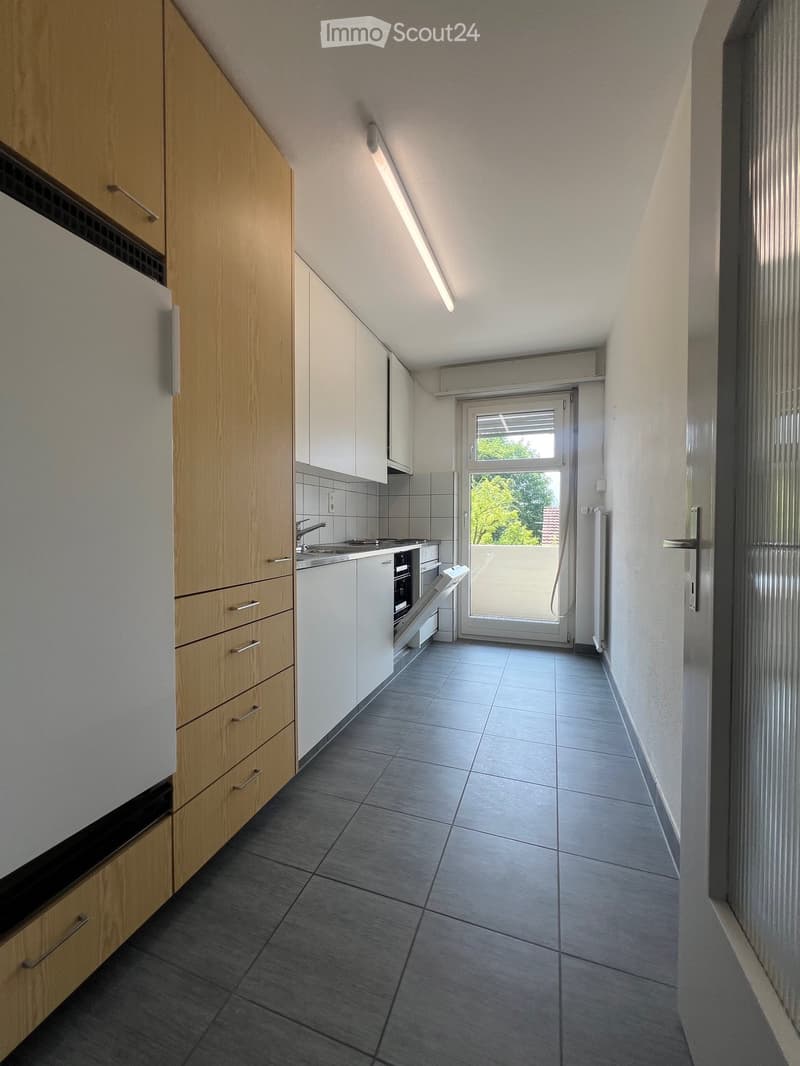 Galley-style kitchen with light wood and white cabinets, tiled floor, and a window overlooking the outdoors. The kitchen includes a built-in oven and dishwasher.
