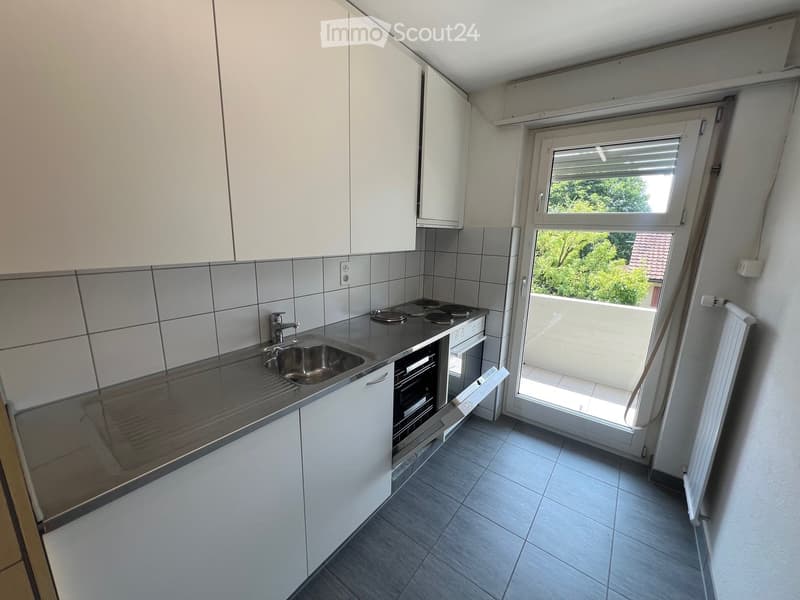 Spacious kitchen with white cabinets, tiled backsplash, stainless steel sink, and a sliding glass door leading to a balcony or terrace with a view of the surrounding greenery.