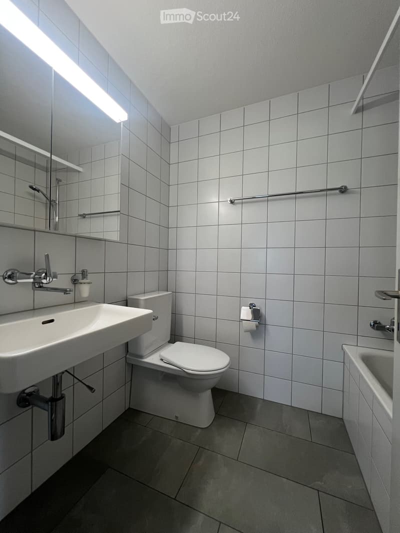 The image shows a modern bathroom with white tiled walls, a sink with a vanity, a toilet, and a shower area. The bathroom appears to be well-lit with a bright, clean aesthetic.