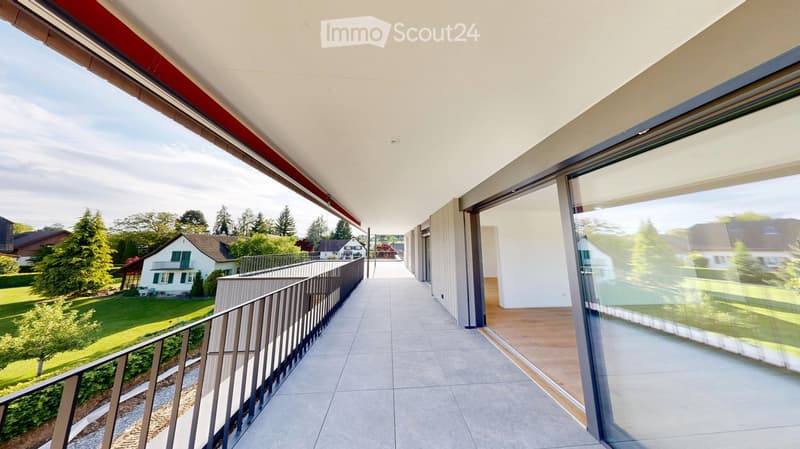 Spacious balcony with modern design, large glass windows, tiled floor, and metal railing, overlooking a green landscape with houses and trees.